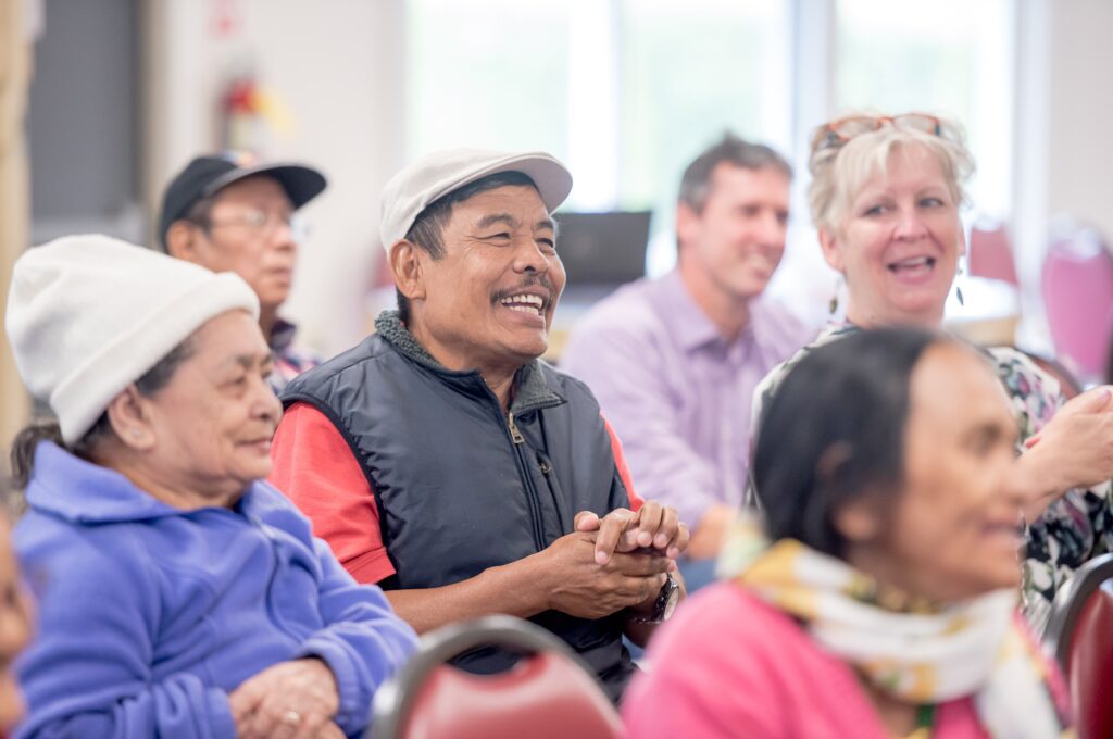 A group of older community members sit in rows of chairs smiling and listening